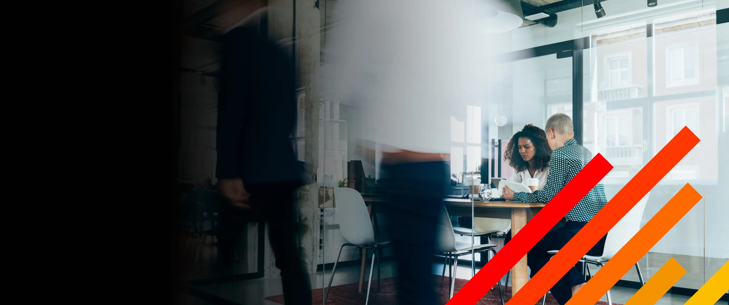 Two female legal professionals working together at a laptop in a glass conference room with colleagues walking by